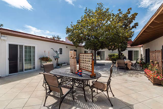 Sunlit courtyard patio with a metal table and chairs, oversized outdoor games, potted plants, and surrounding single-story building entrances.