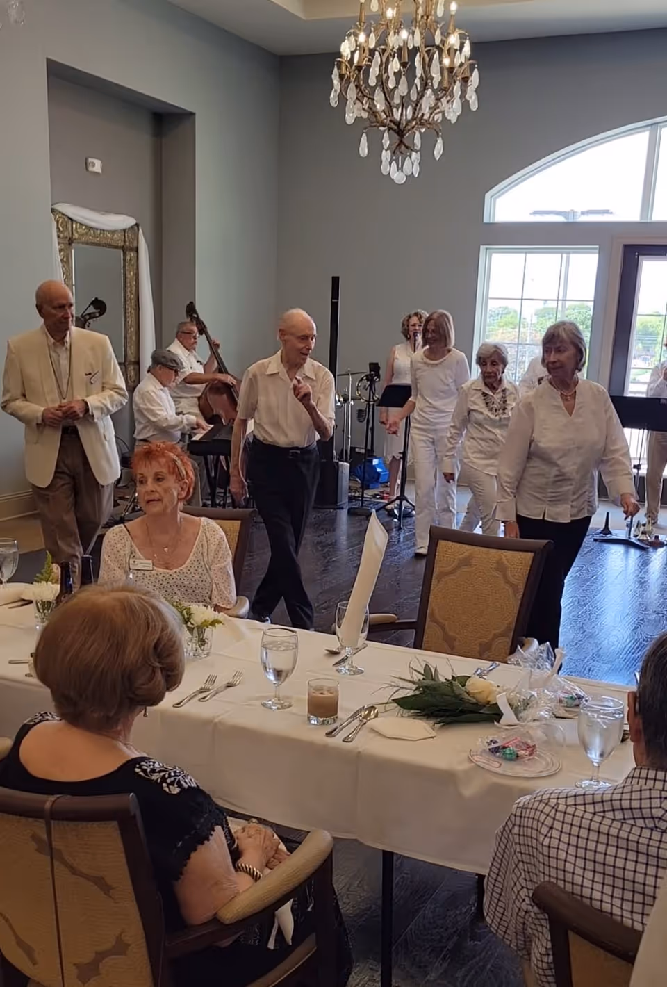 A group of elderly people in a dining room setting at Franklin Park Alamo Heights. Some individuals are seated at a table set with glasses, utensils, and flowers, while others are standing or walking near a live band playing instruments in the background. The room features a chandelier and large windows letting in natural light.