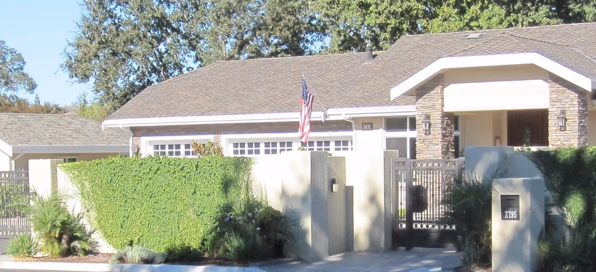 Exterior view of a residential-style building with a gated entrance, stone pillars, and a well-maintained hedge. An American flag is mounted on the roof near the entrance.