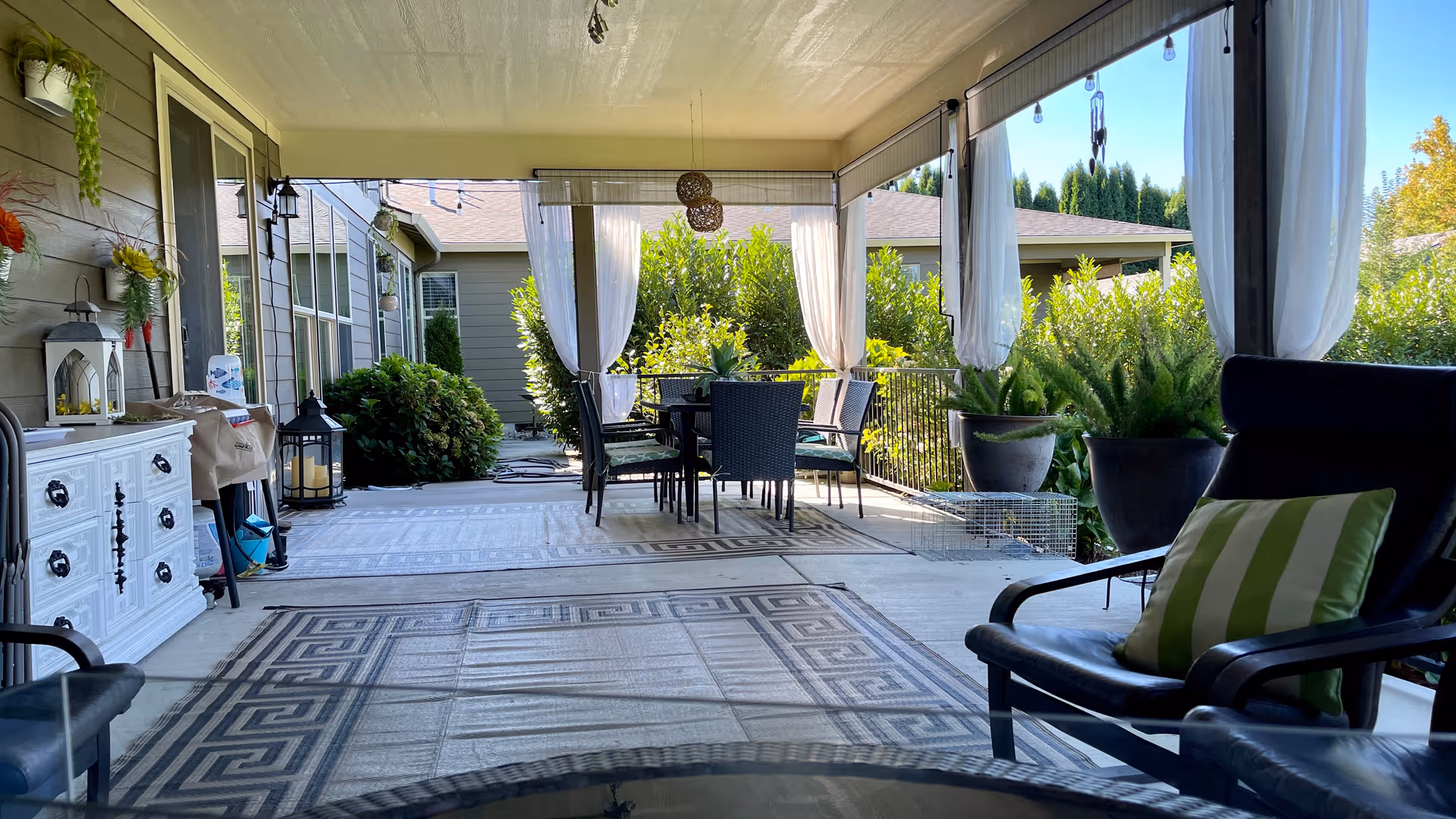 Covered outdoor patio with seating, a dining table, potted plants and decorative curtains.