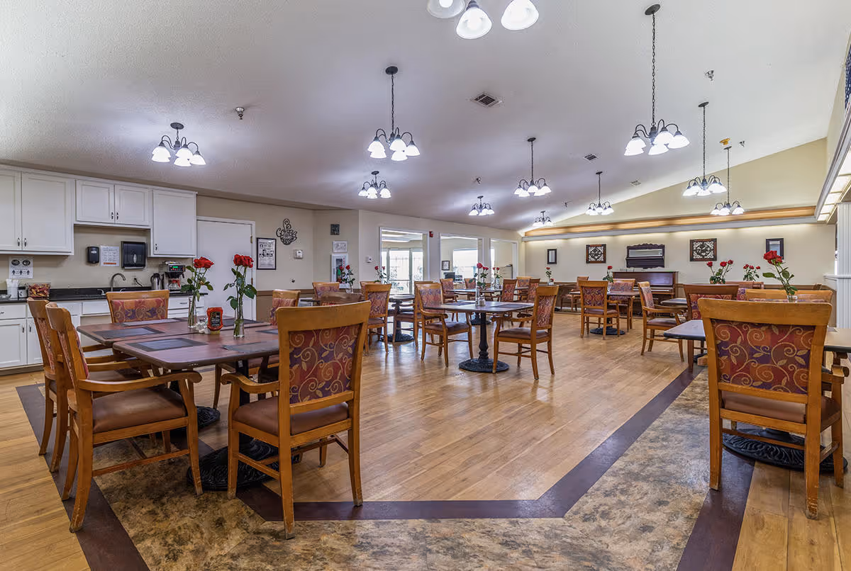 A spacious dining room with multiple wooden tables and chairs arranged neatly. Each table has a vase with red roses. The room has wooden flooring with a decorative border, white cabinets and a countertop along one wall, and multiple ceiling lights providing bright illumination.