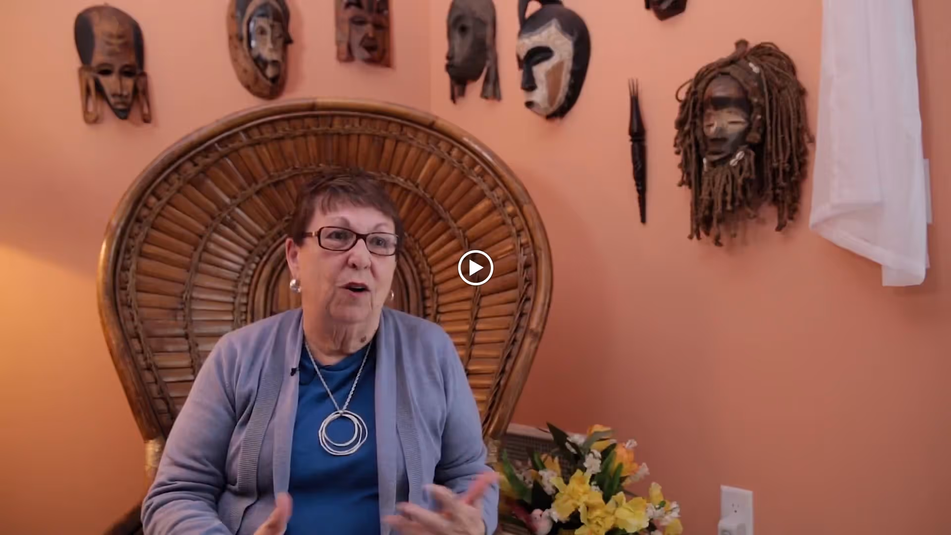 An elderly woman wearing glasses, a blue shirt, and a gray cardigan is sitting in a large wicker chair with a rounded back. Behind her on the peach-colored wall are several decorative African-style masks. There is a bouquet of yellow and white flowers on a table next to her.