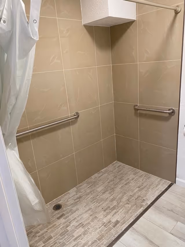 A walk-in shower area with beige tiled walls and a textured tile floor. There are two stainless steel grab bars mounted on the walls for support, and a white shower curtain partially pulled to the side.