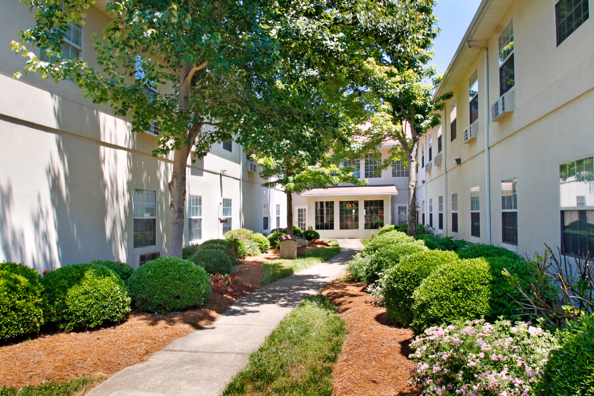 Outdoor courtyard area between two beige buildings with multiple windows and air conditioning units. The courtyard features a concrete walkway, green bushes, trees providing shade, and some flowering plants under a clear blue sky.