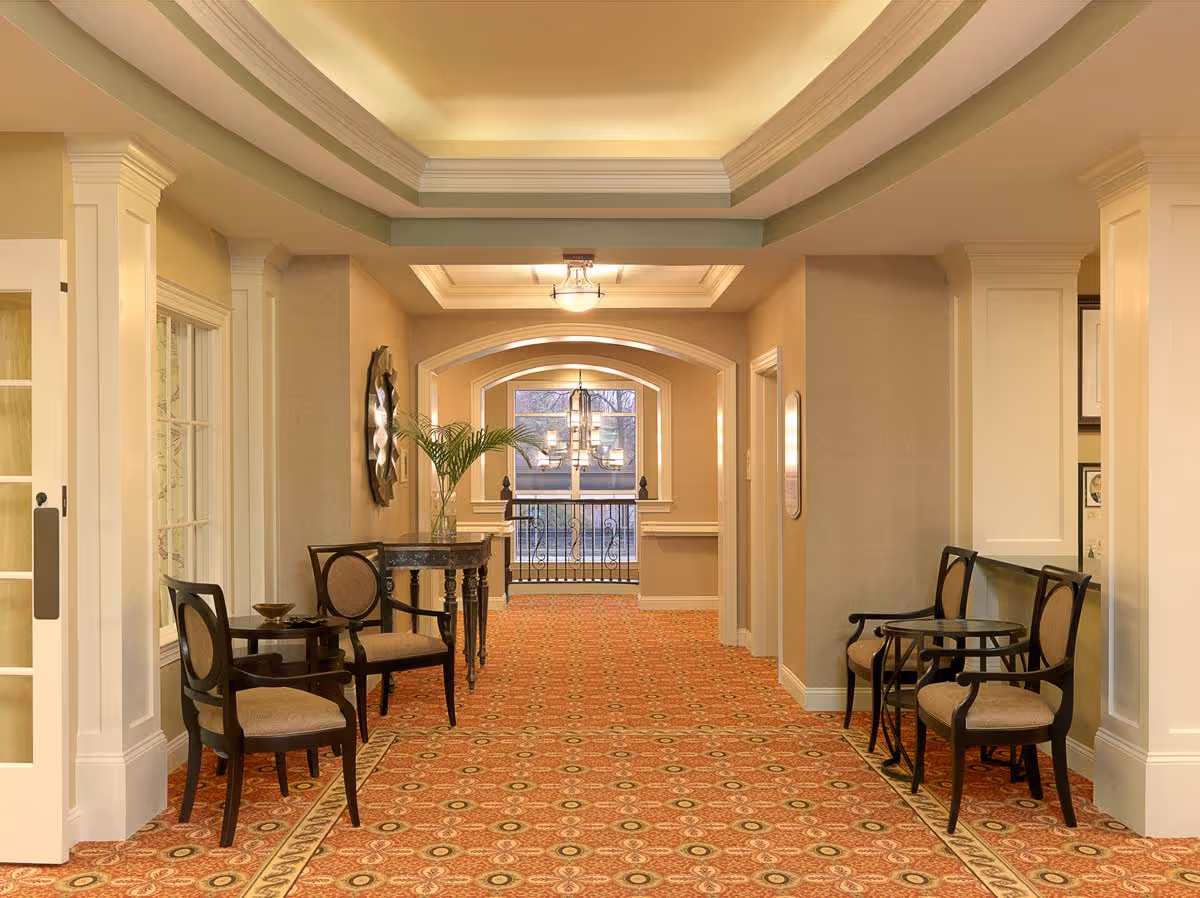 A well-lit hallway in a senior living facility with patterned carpet, beige walls, and white trim. The hallway features several dark wood chairs with beige cushions arranged in pairs along the walls, small tables, a decorative mirror, and a plant on a console table. At the end of the hallway, there is a window with a view outside and a hanging light fixture.