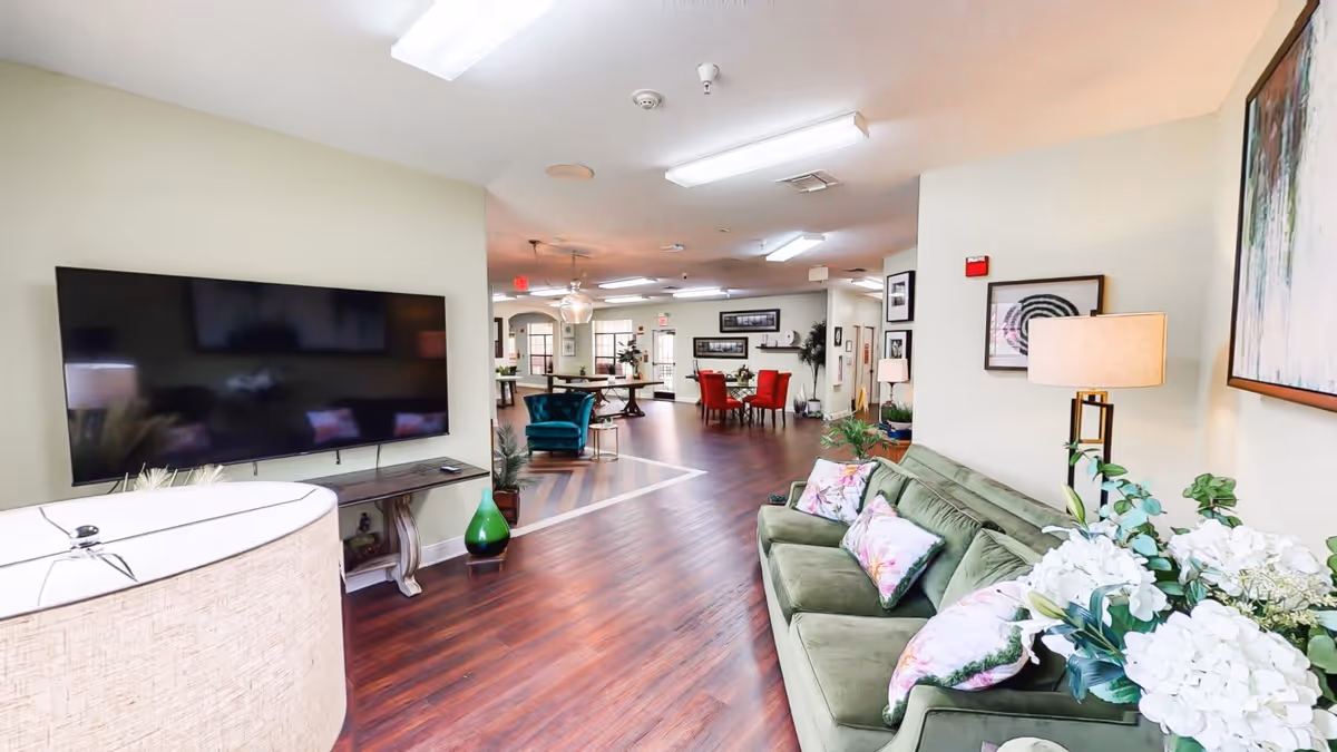 Interior view of a senior living facility lounge area with a green sofa adorned with floral pillows, a large flat-screen TV mounted on the wall, wooden flooring, and various chairs and tables arranged in the background under bright ceiling lights.