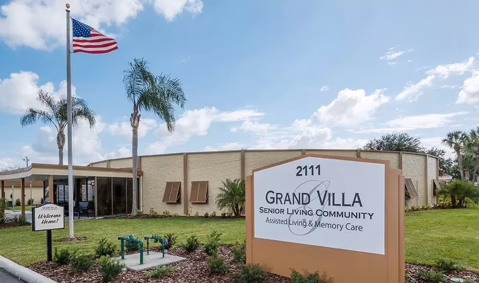 Exterior view of Grand Villa Senior Living Community building with a large sign displaying the name and address 2111. The building is surrounded by green grass, palm trees, and an American flag on a flagpole. A smaller sign near the entrance reads 'Welcome Home!'.