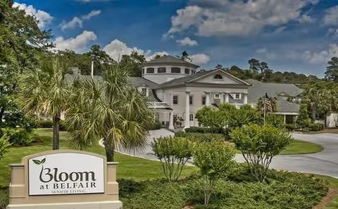Exterior view of Bloom At Belfair senior living facility showing a large white building with columns, surrounded by greenery and palm trees under a partly cloudy sky. A sign in the foreground reads 'Bloom at Belfair Senior Living'.