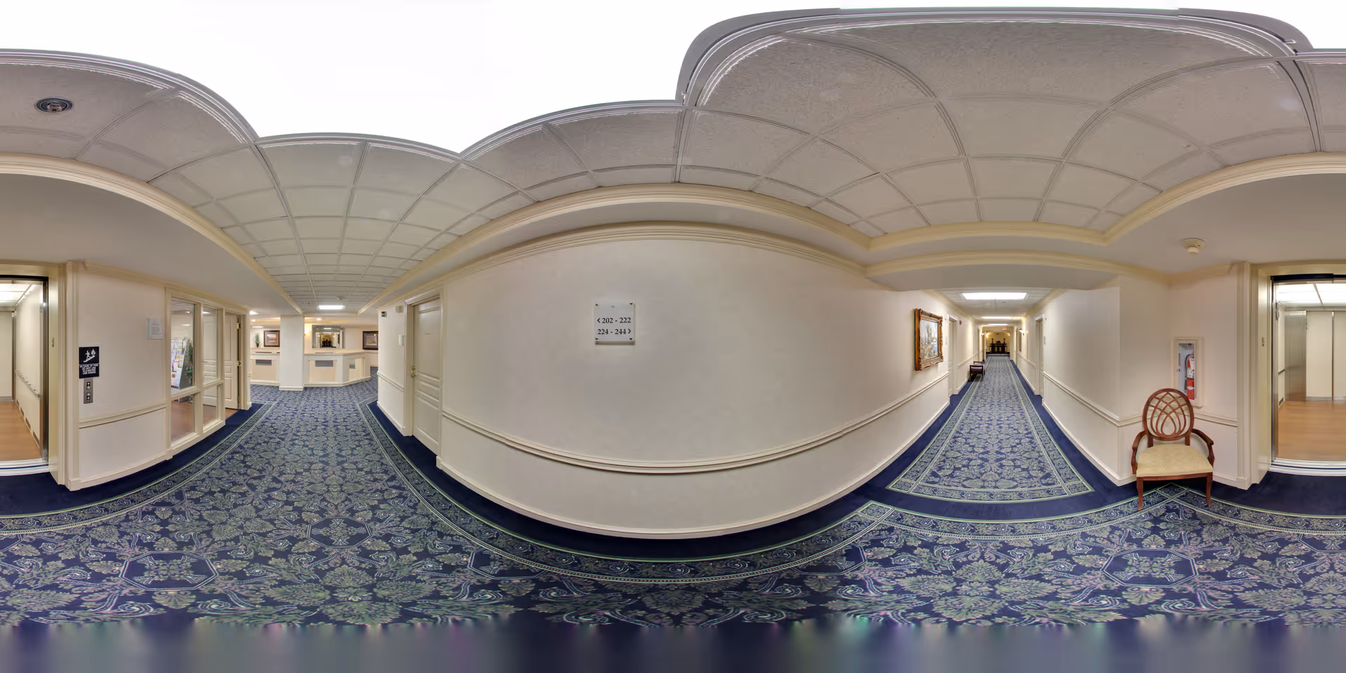 A long, carpeted hallway in a senior living facility with patterned blue and green carpet, cream-colored walls, and a drop ceiling with fluorescent lighting. There are doors along the hallway, a chair with a round backrest on the right side, a fire extinguisher mounted on the wall, and a framed picture hanging on the wall. The hallway leads to a reception or common area with more lighting and furniture visible in the distance.