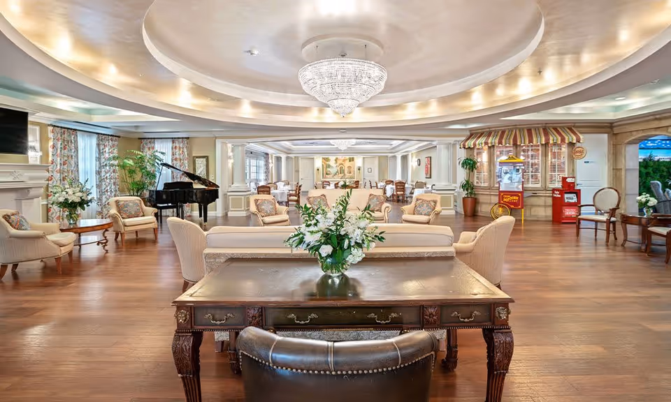 Spacious and elegant senior living facility common area with wooden flooring, comfortable armchairs and sofas, a grand piano, floral arrangements, a chandelier, and a popcorn machine under a striped awning. The room is well-lit with natural light from large windows and has a cozy, inviting atmosphere.