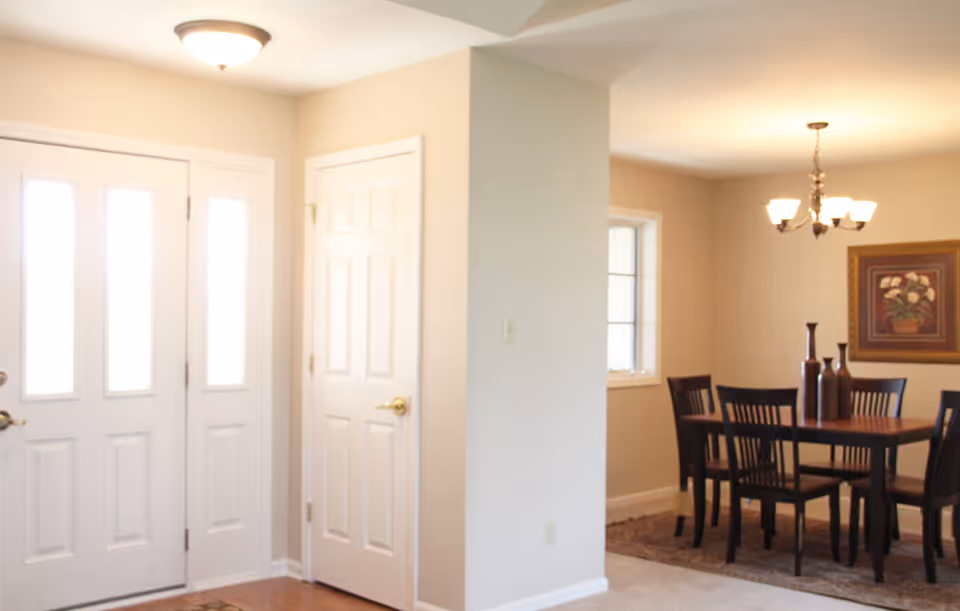 Interior view of a residential space showing an entryway with a white front door and a closet door. Adjacent to the entryway is a dining area with a wooden dining table, six chairs, a chandelier overhead, and a framed floral painting on the wall.