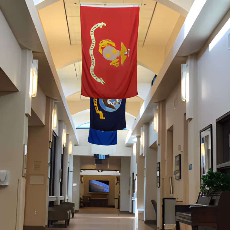 Interior hallway of the William E. Christoffersen Salt Lake Veterans Home with high ceilings and multiple flags hanging from above. The hallway is lined with windows, wall-mounted lights, and seating areas along the sides.