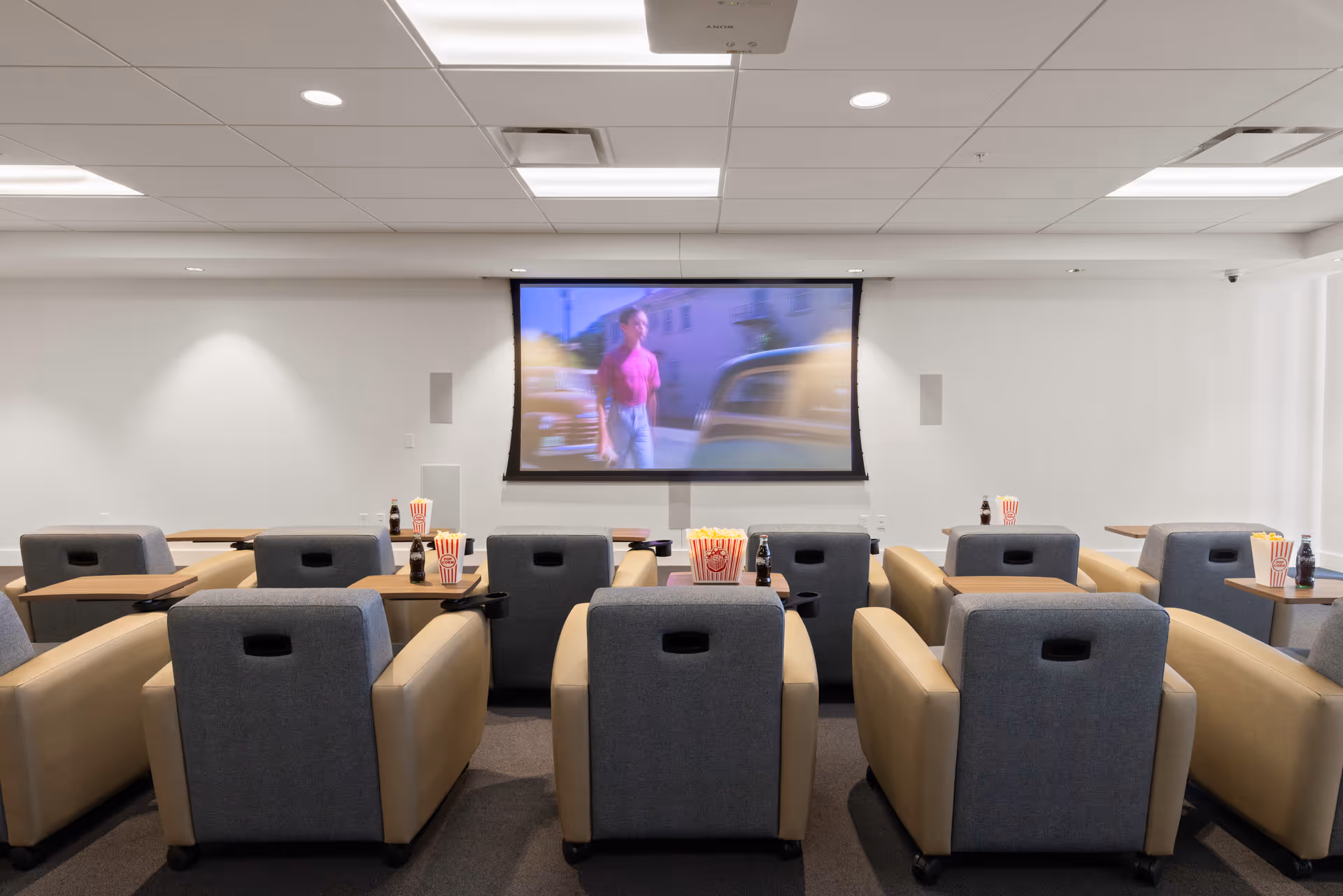 A small theater room with multiple rows of comfortable armchairs facing a large screen showing a movie scene. Each chair has a small table attached, holding popcorn containers and soda bottles. The room has a white ceiling with recessed lighting and a projector mounted above.
