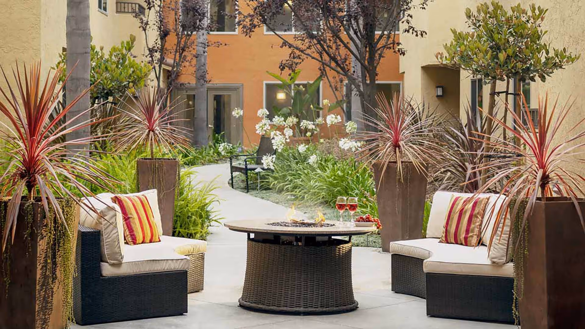 Outdoor seating area with cushioned chairs and a round fire pit table surrounded by tall planters with spiky plants. The area is part of a courtyard with a pathway, greenery, and buildings with windows in the background.