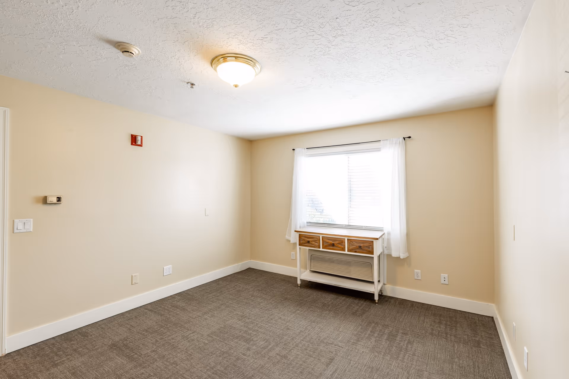 Empty carpeted bedroom with beige walls, a window with sheer curtains and a small console table over a wall heater.