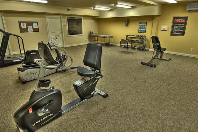 A fitness center room with exercise equipment including a recumbent bike, a treadmill, a weight bench, dumbbells on a rack, and a table with a chair. The room has beige walls and carpeted floor, with fluorescent lighting on the ceiling. There are posters on the walls including one with fitness center rules.