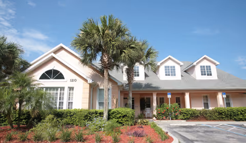 Exterior view of a single-story building with a light peach facade, gray roof, and several windows. The building is surrounded by green shrubs, palm trees, and a small garden area. There is a parking lot in front with marked handicap parking spaces under a clear blue sky.