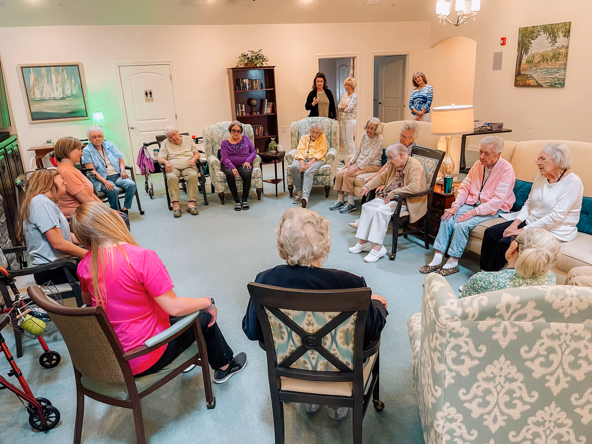 A group of elderly residents and caregivers seated in a circle in a communal living room of an assisted living facility.