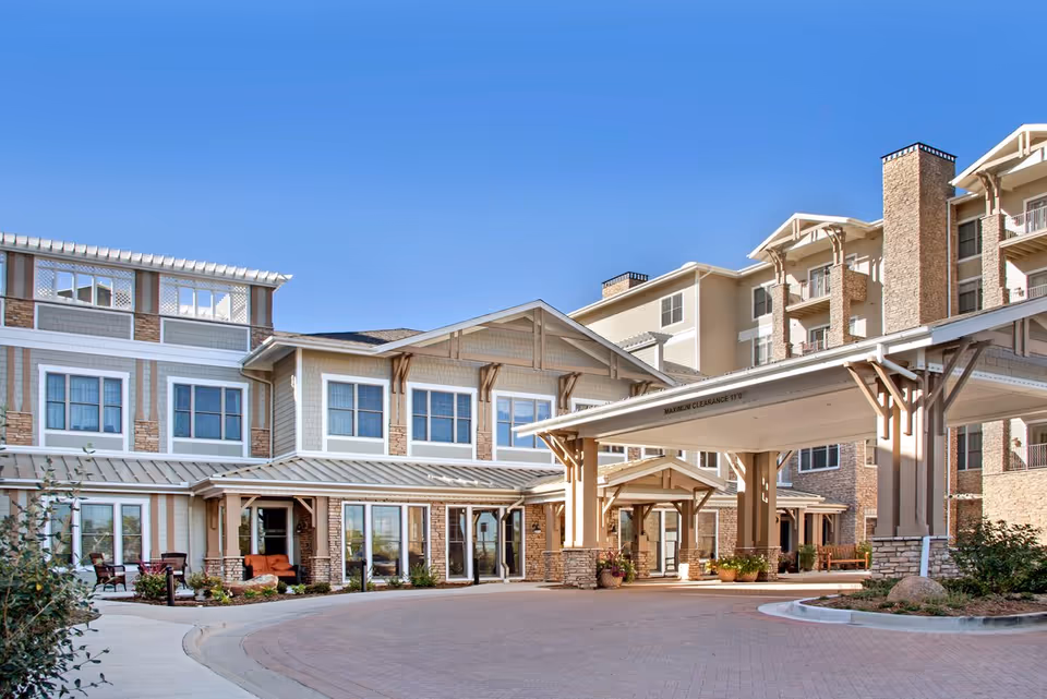 Front exterior of a multi-story senior living facility with a covered porte-cochere entrance, walkway, and landscaping under a clear blue sky.