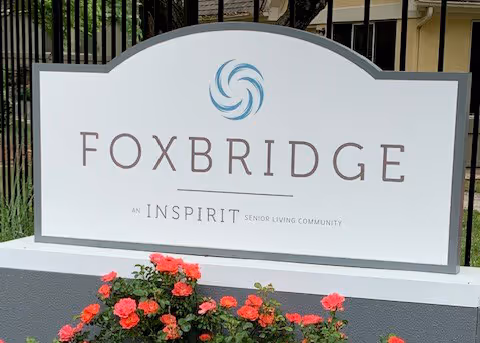 A white entrance sign with a blue swirl logo reading "FOXBRIDGE" in front of a fence and red flowers.