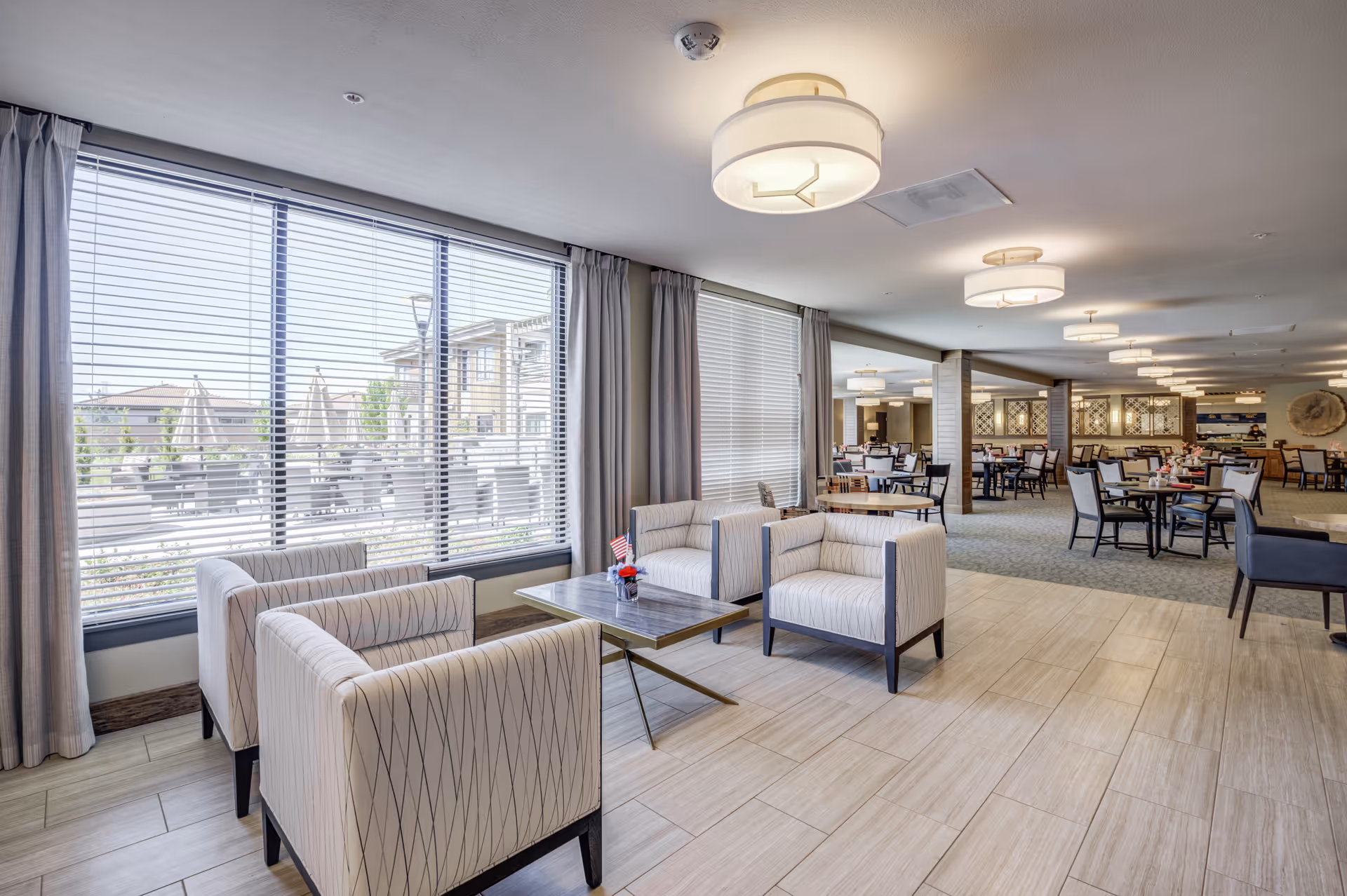 A spacious and well-lit common area in a senior living facility featuring a seating arrangement with four cushioned armchairs around a small rectangular table near large windows with blinds and curtains. In the background, there are multiple dining tables and chairs arranged neatly under modern ceiling lights.
