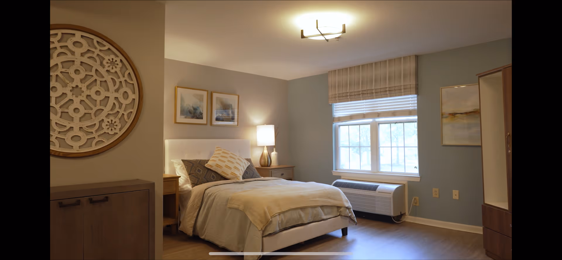 Well-lit decorated bedroom with a bed, nightstand and lamp, window with a roman shade, and decorative wall art.