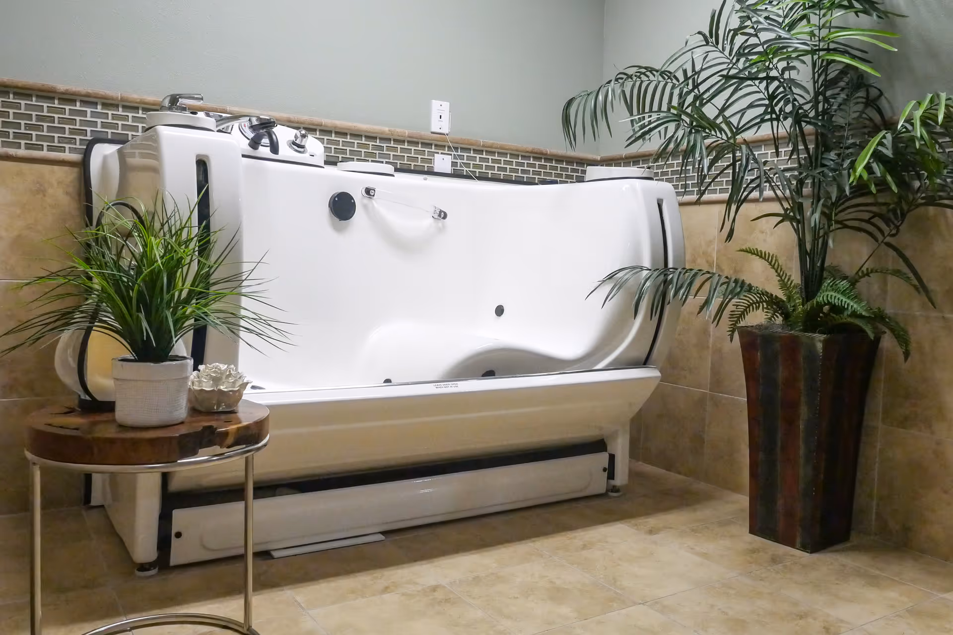 A white walk-in bathtub with safety handles and controls, placed against a tiled wall with a decorative tile border. There is a small round wooden table with a potted plant and decorative item on the left side of the tub, and a tall potted plant on the right side. The floor is tiled in a beige color.