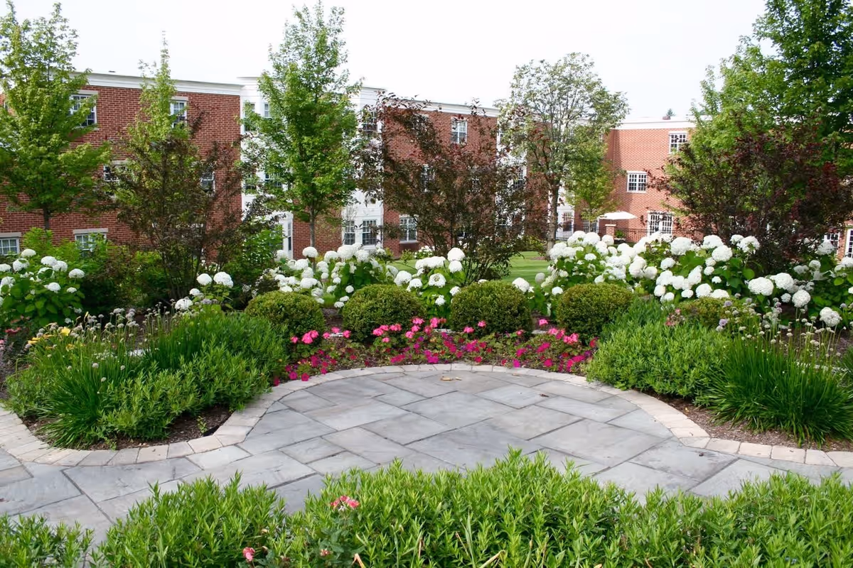 A landscaped garden area with a stone-paved circular patio surrounded by green shrubs, white hydrangeas, pink flowers, and trees. In the background, there is a red brick building with multiple windows.