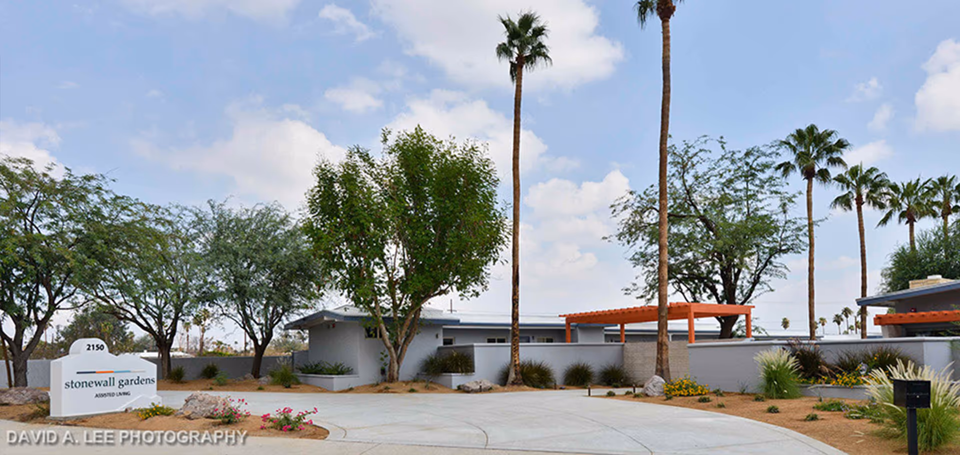 Exterior view of Stonewall Gardens Assisted Living facility showing a driveway, landscaped area with trees and plants, and a sign with the facility name and address 2150. The sky is partly cloudy.