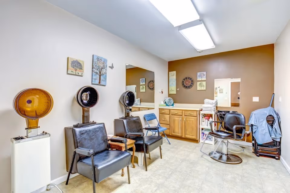 Interior view of a hair salon area with three black salon chairs, two with hair dryers above them. There is a wooden cabinet with drawers and shelves holding towels and supplies against a brown accent wall. The room has light-colored tiled flooring and white walls with decorative wall art and a clock.