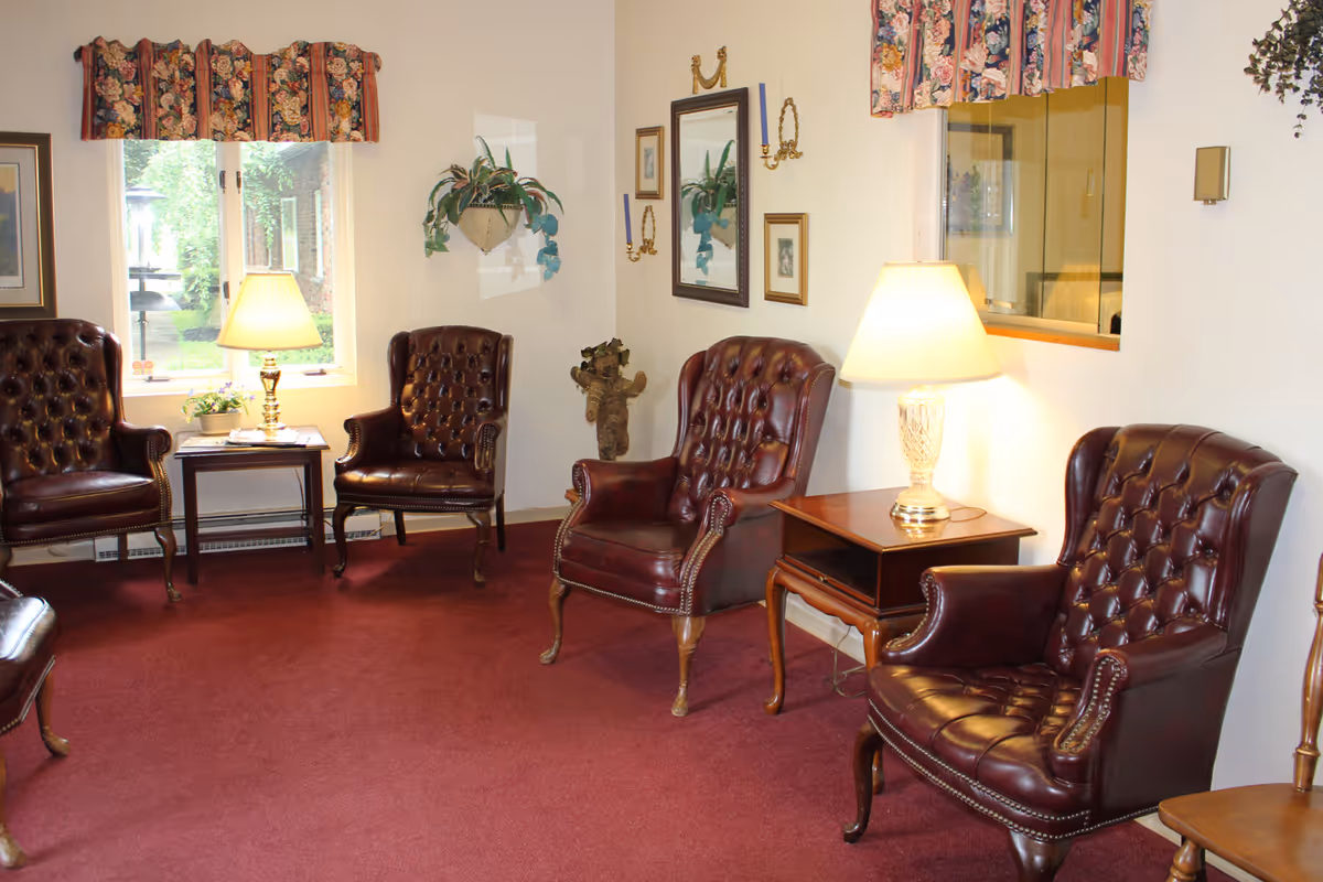 A cozy living room with four dark brown leather tufted armchairs arranged around two wooden side tables with lamps. The room has a red carpet, cream-colored walls, floral valances on the windows, framed pictures on the walls, and a hanging plant.