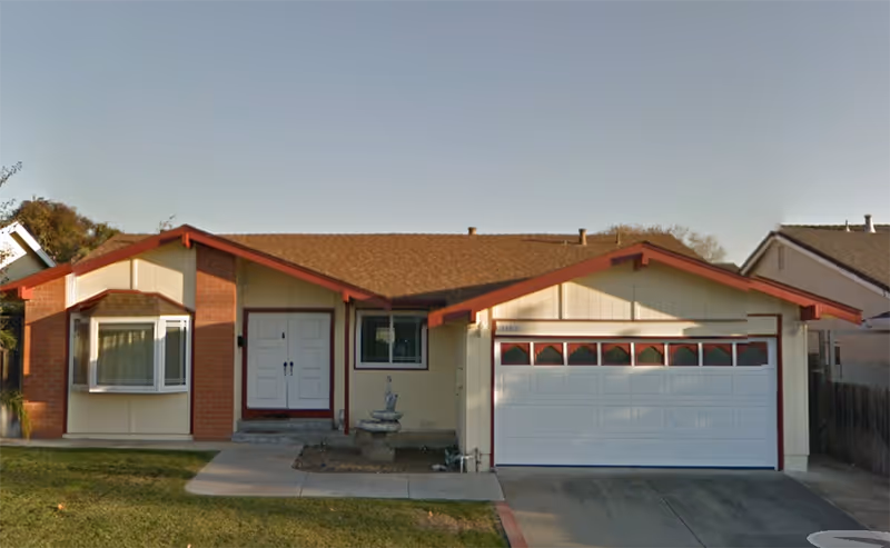 Single-story residential building with a brown shingled roof, beige siding, and red trim. The house features a white double front door, a bay window on the left, a two-car garage with a white door on the right, and a small fountain in front of the house. There is a concrete driveway and a grassy lawn in front.