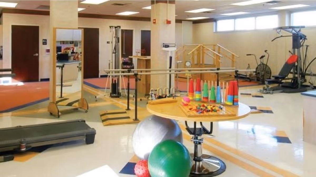 A spacious physical therapy or rehabilitation room with exercise equipment including a treadmill, parallel bars, balance balls, a mirror, and various therapy tools on a round table. The room has large windows allowing natural light and a clean, organized layout.