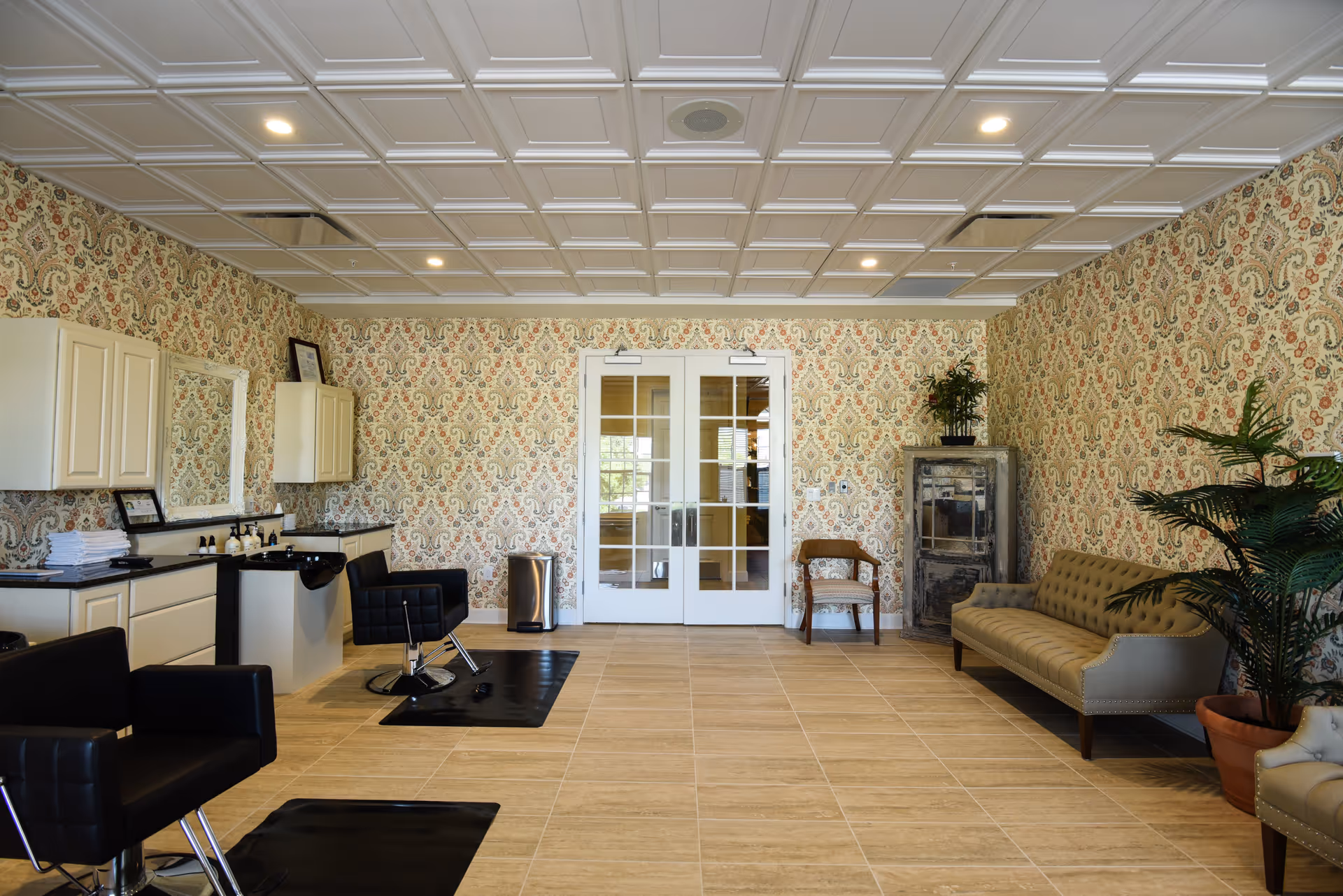 Interior of a salon area with two black salon chairs in front of a counter with sinks and mirrors. The room has patterned wallpaper, a tiled floor, a beige tufted sofa, a wooden chair, a tall cabinet, and potted plants. Double glass doors are visible at the back.