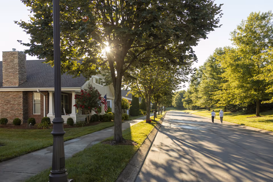 A peaceful residential street in a senior living community with houses lined with trees and a sidewalk. Two people are walking down the road on a sunny day with the sun shining through the tree leaves.