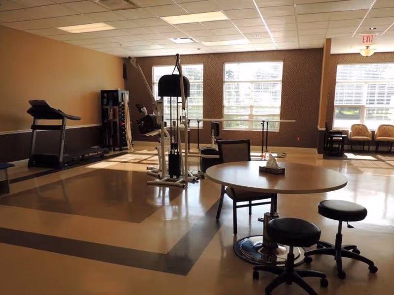 A spacious room with exercise equipment including a treadmill and a multi-station gym machine, a round table with chairs and stools, large windows allowing natural light, and a seating area with armchairs near the windows.