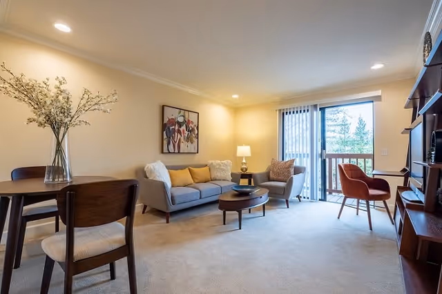 A cozy living room with a gray sofa adorned with yellow and white pillows, a matching armchair, a round wooden coffee table, and a side table with a lamp. There is a wooden dining table with chairs and a vase with flowers on it. A sliding glass door with vertical blinds opens to a balcony with a view of trees. The room has beige walls and carpet, and a colorful abstract painting hangs above the sofa.
