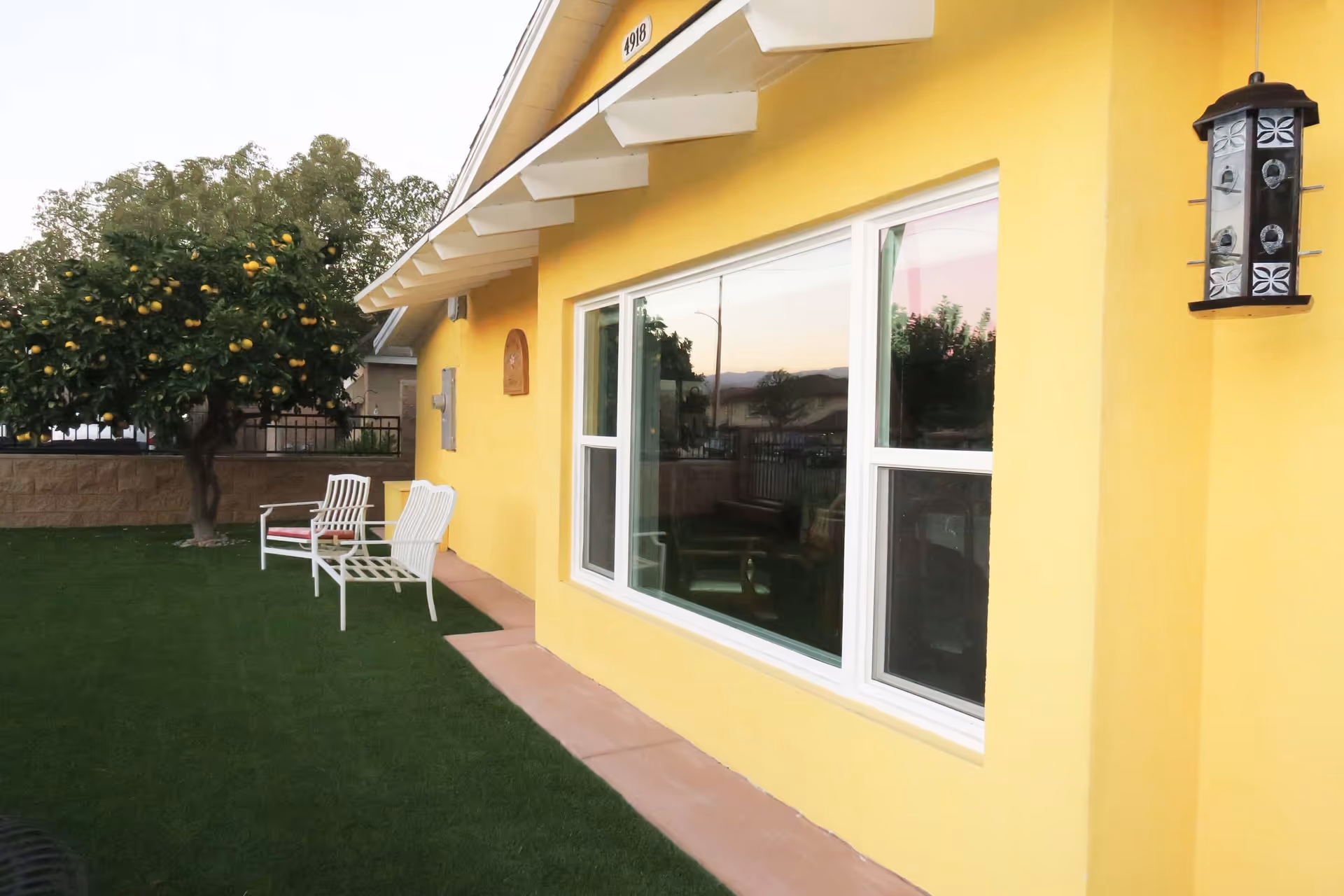 Exterior view of a yellow building with a large window, two white chairs with red cushions on a green lawn, a lemon tree with fruit, and a decorative black lantern mounted on the wall.