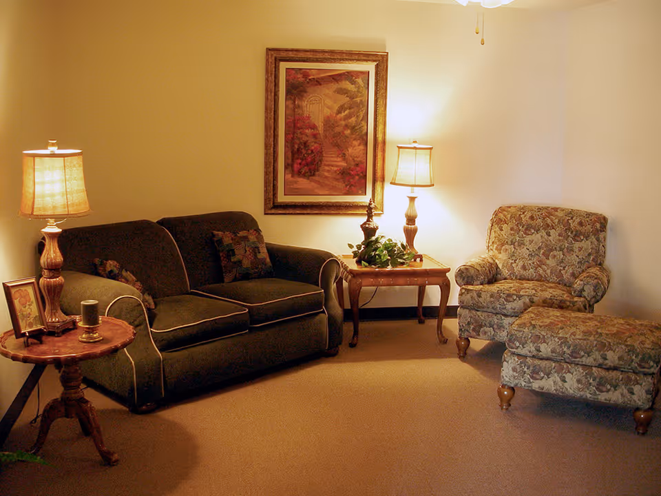 A cozy living room with a dark green sofa, a floral armchair with matching ottoman, two wooden side tables each with a lamp, a framed painting on the wall, and a small framed picture on one of the tables.