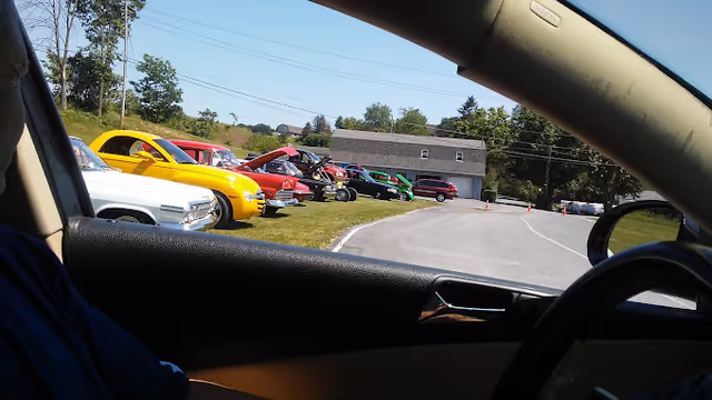 View from inside a car looking out through the passenger side window at a row of colorful vintage cars parked on a grassy area next to a curved road, with trees and a building in the background under a clear sky.