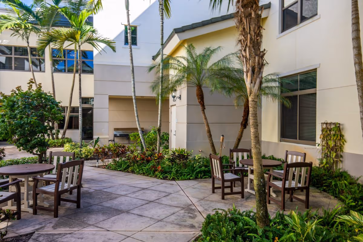 Outdoor courtyard area at Luxe Senior Living at Wellington featuring round wooden tables with chairs, surrounded by palm trees, plants, and shrubs, adjacent to a building with windows.
