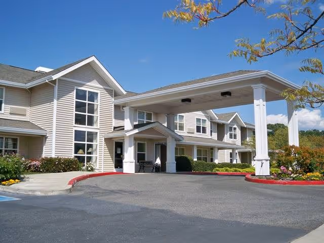 Front entrance of a two-story beige senior living building with a covered porte-cochère and landscaped grounds under a blue sky.