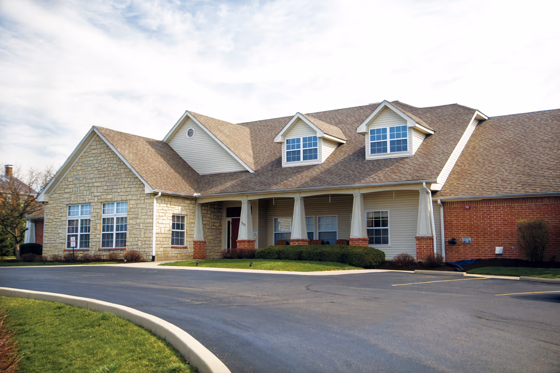Front exterior of a single-story senior living building with a covered porch, dormer windows, and an adjacent parking area.