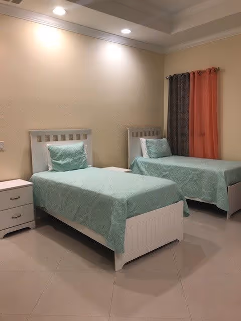 A bedroom with two single beds featuring white wooden frames and light green bedding. Each bed has a matching pillow. There is a white nightstand with two drawers between the beds. The walls are light beige, and there are two sets of curtains, one dark brown and one orange, hanging on the window. The room is well-lit with recessed ceiling lights.