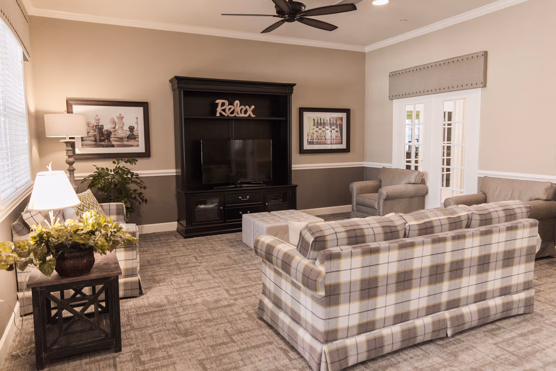 A cozy living room with plaid sofas and armchairs arranged around a black entertainment center with a TV and a decorative 'Relax' sign. The room features framed chess-themed artwork on the walls, a ceiling fan, carpeted floor, and a side table with a lamp and a plant.