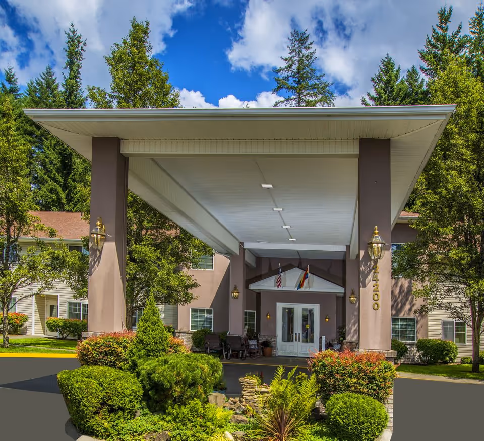 Entrance of Fountain Court Senior Living facility with a covered driveway, surrounded by well-maintained greenery and trees under a blue sky with some clouds.