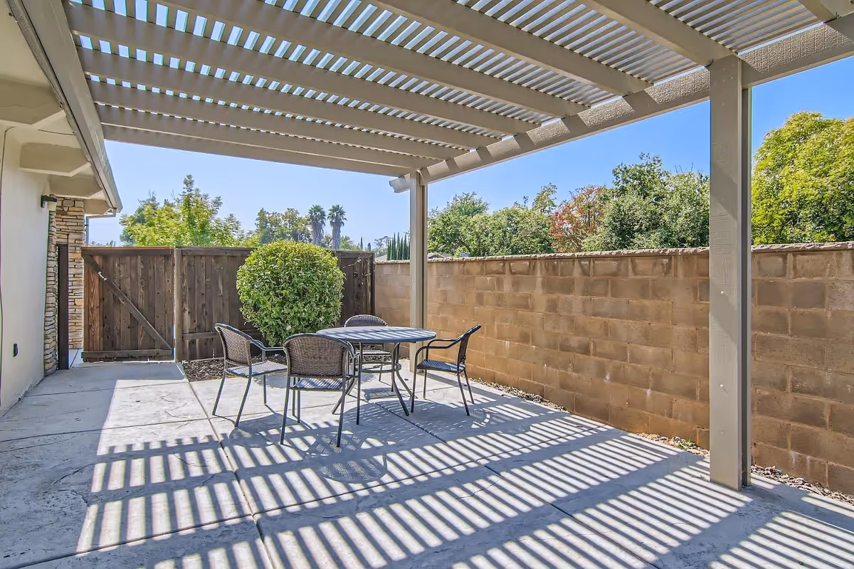 Outdoor covered patio with a pergola casting striped shadows over a table and four chairs next to a brick wall and wooden gate.