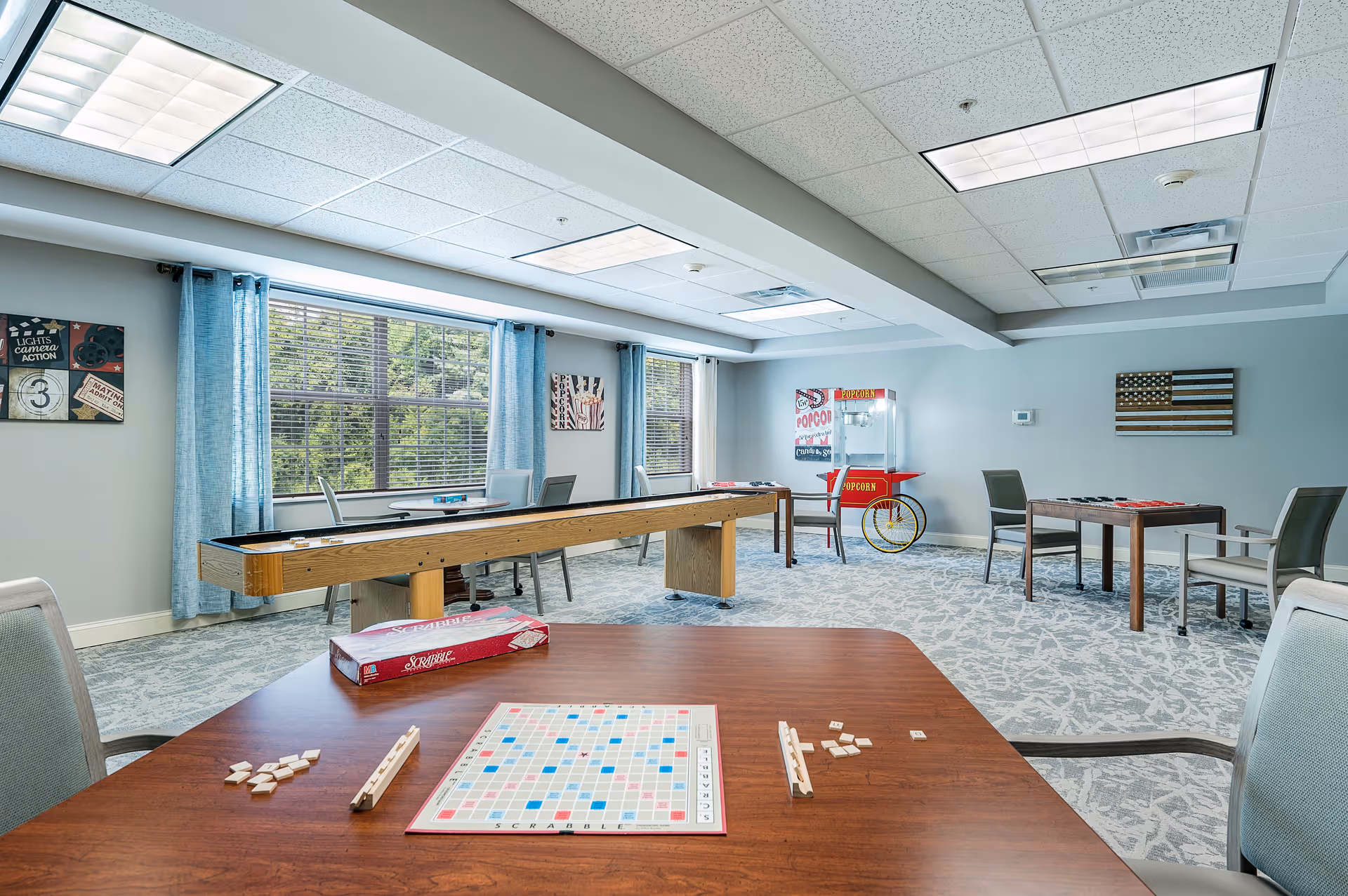 A bright and spacious activity room with large windows covered by blue curtains. The room features a shuffleboard table, a popcorn machine, and several tables with chairs. One table in the foreground has a Scrabble board game set up with tiles and racks. The walls are decorated with framed artwork, including an American flag and movie-themed prints.