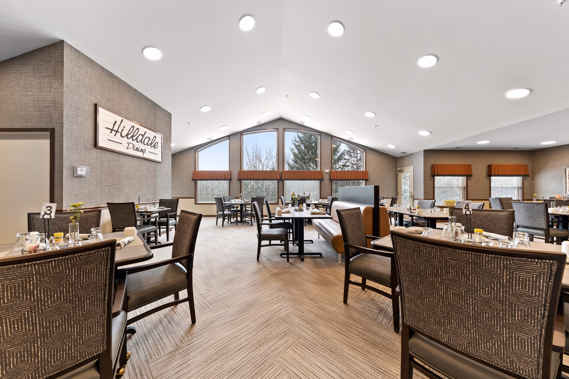 Bright senior dining room with tables and chairs arranged around a central walkway and a 'Hilldale Dining' sign on the left wall.