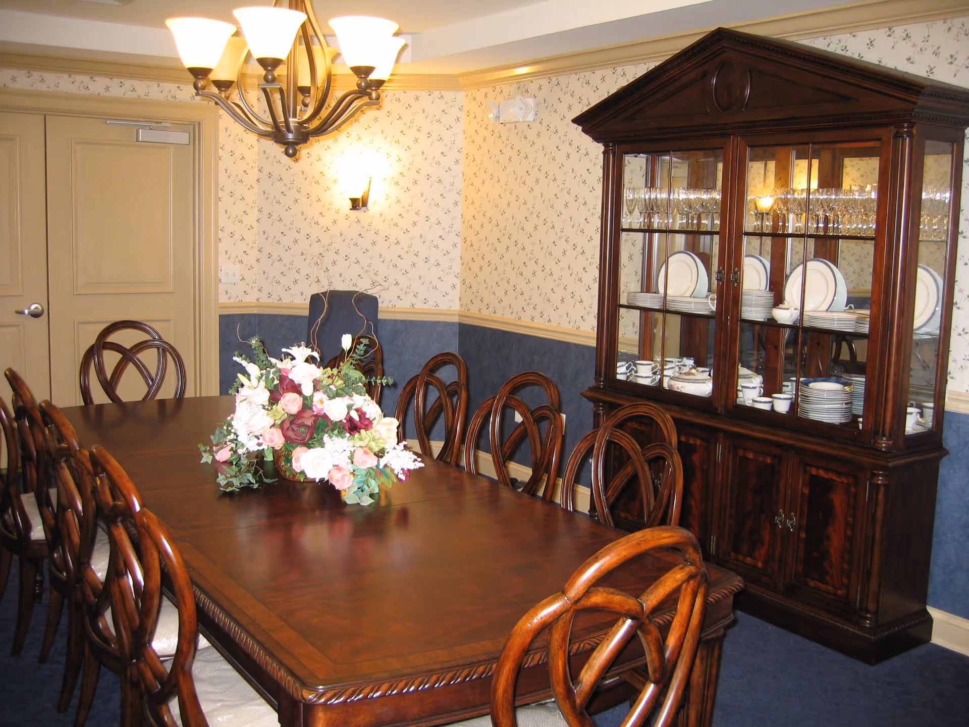 A formal dining room with a long wooden table surrounded by wooden chairs with cushioned seats. A floral centerpiece is placed in the middle of the table. On the right side, there is a large wooden china cabinet displaying plates, cups, and glassware. The walls have floral wallpaper with a chair rail separating a blue lower section. A chandelier with multiple lights hangs above the table, and a wall sconce provides additional lighting.